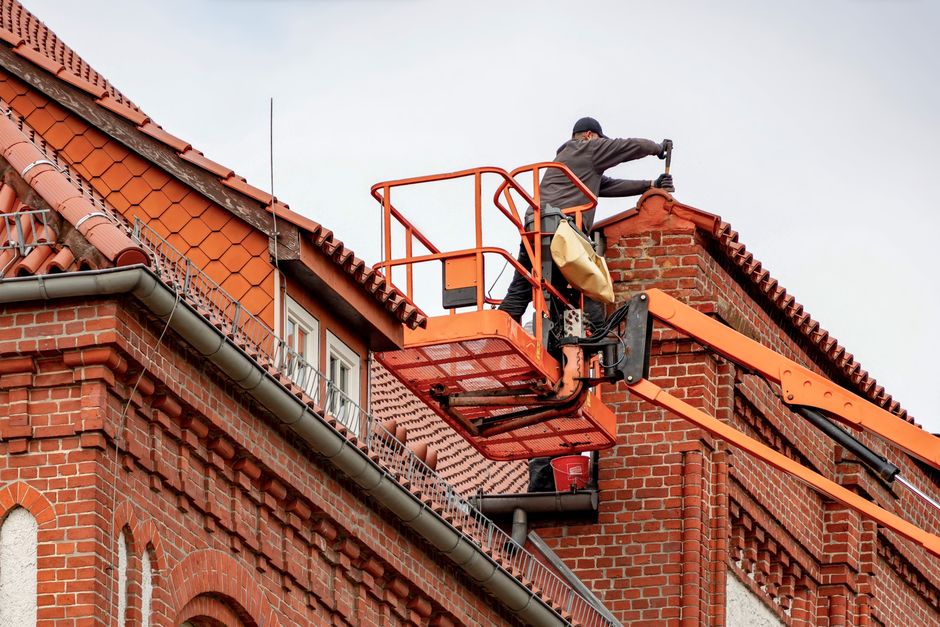 A professional roofer in safety gear diligently installing new shingles on a suburban home's roof under a clear blue sky.