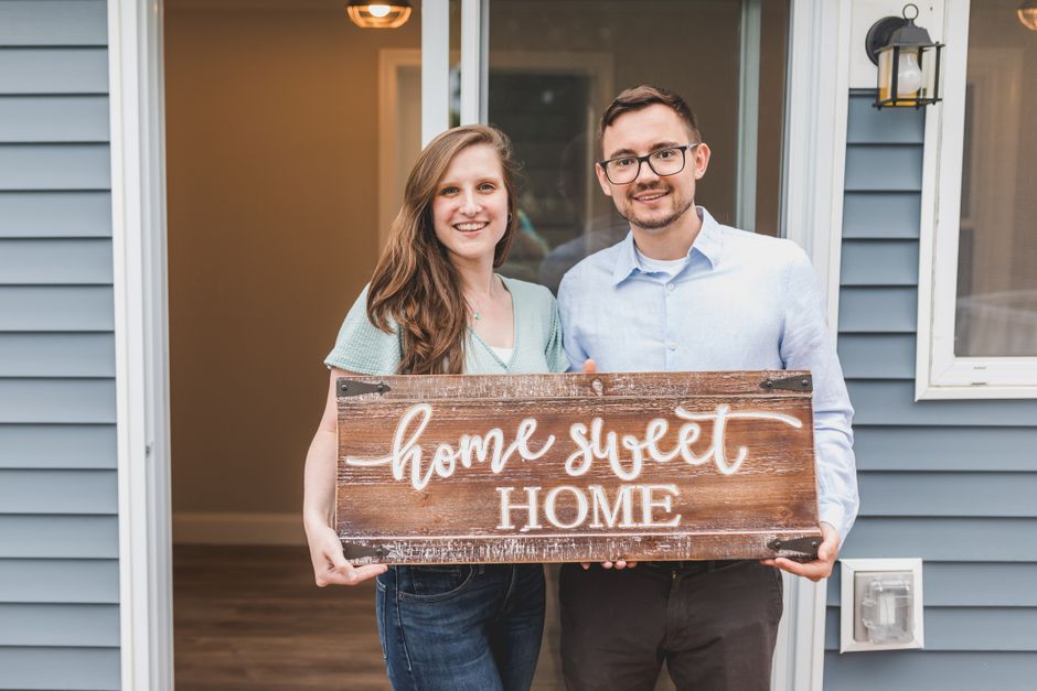 A smiling couple stands on the front porch of a charming house, holding up a set of new house keys, symbolizing a successful home purchase.